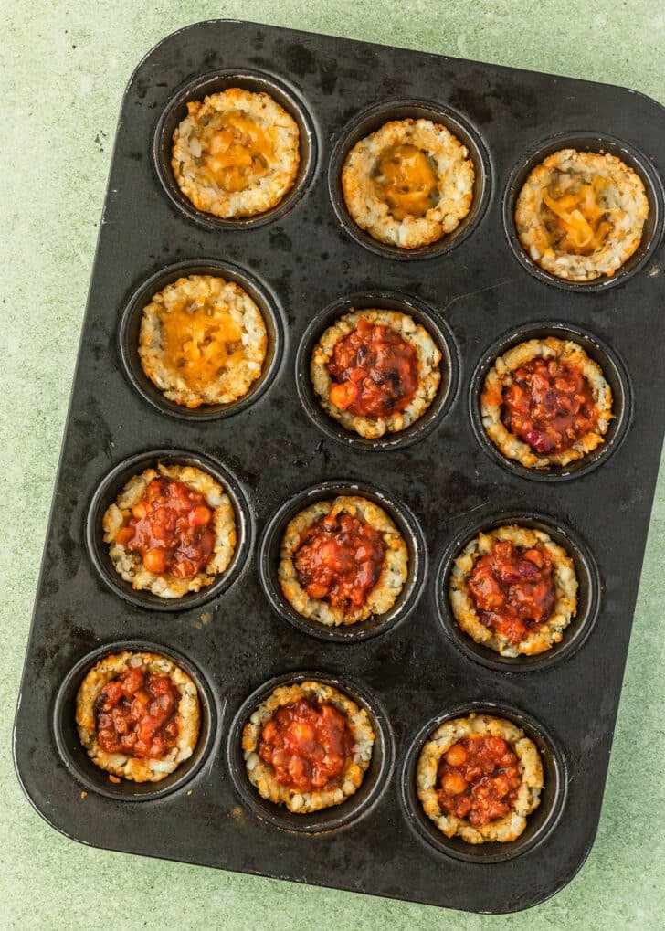 A cupcake pan of potato cups with chili on a green counter.