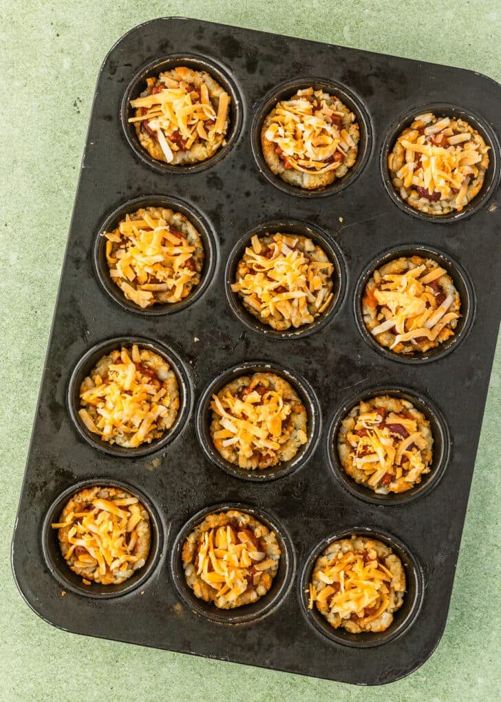 A cupcake pan of tater tot chili cheese cups on a light green table.