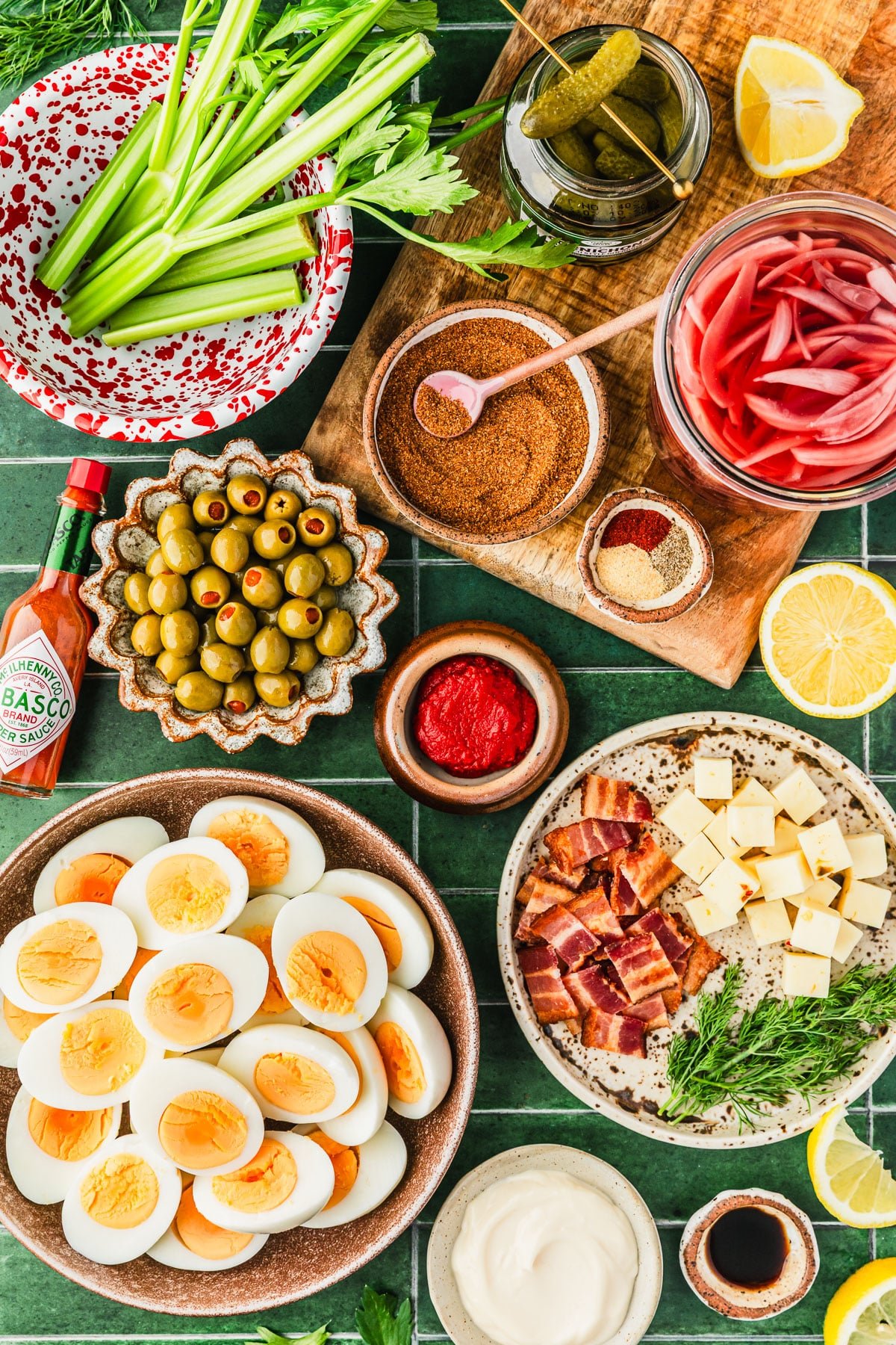 Brown bowls of hard-boiled eggs, bacon, cheese, dill, olives, tomato paste, Cajun seasoning, and spices next to a red and white bowl of celery, and jars of pickles and red onions on a green tiled counter.