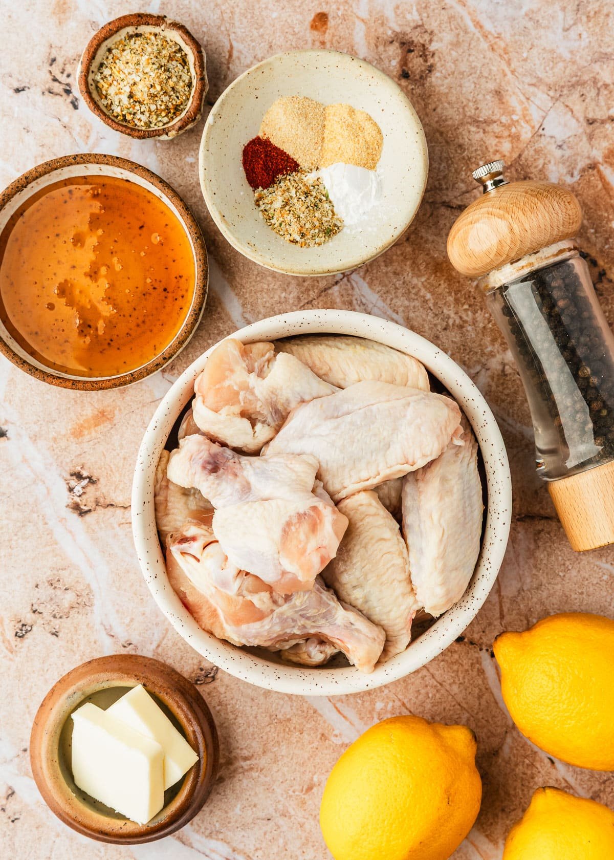 Brown and white bowls of chicken, honey, spices, and butter next to a pepper grinder and lemons.