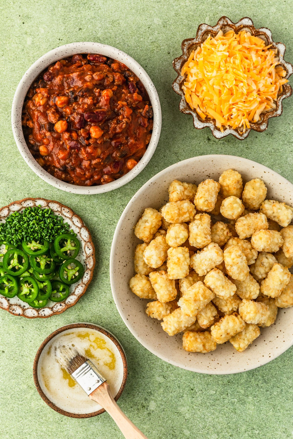 White and brown bowls of chili, tater tots, shredded cheddar, jalapenos and chives, and melted butter on a green counter.