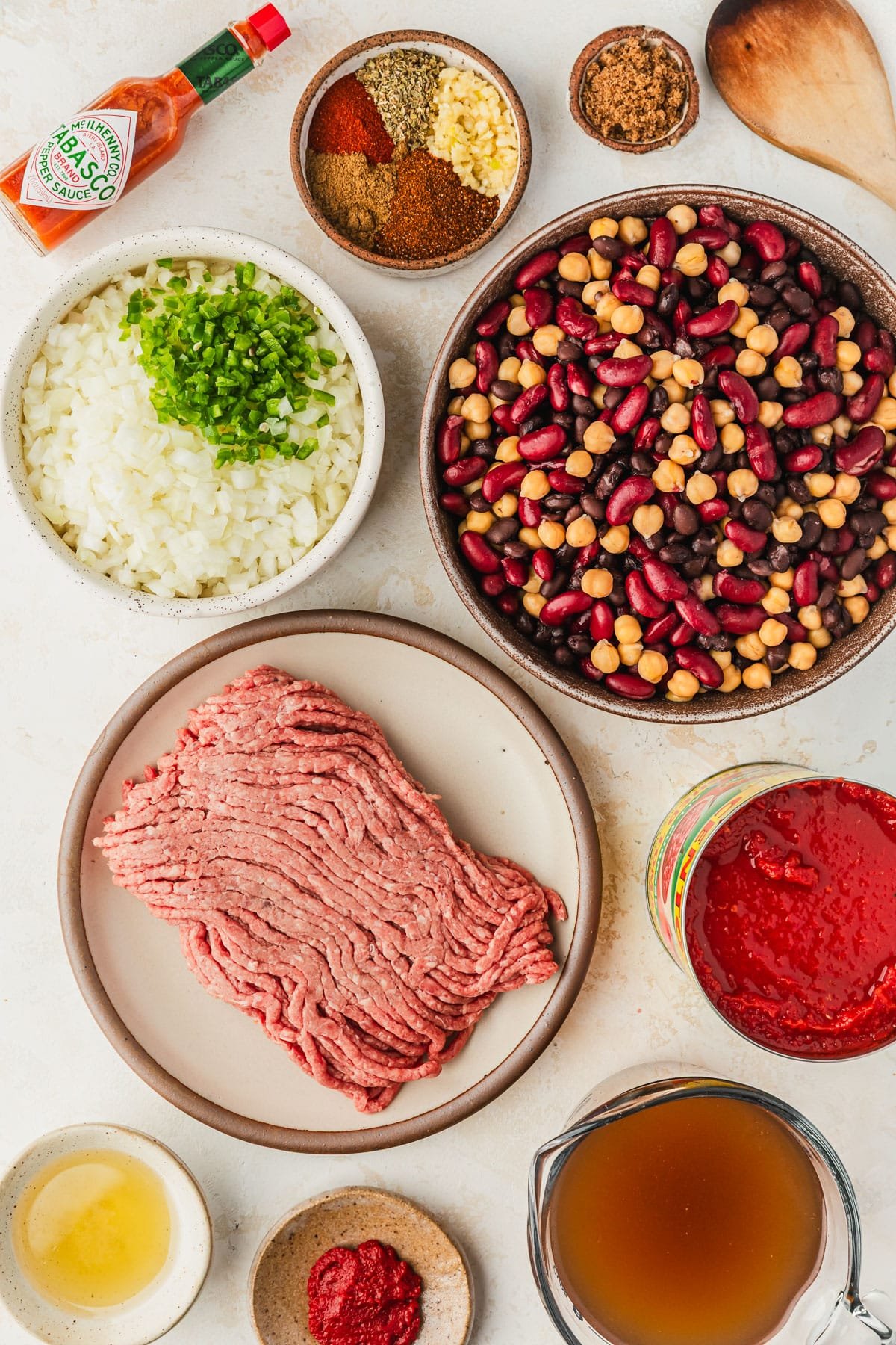 Brown and white bowls of beans, meat, veggies, tomato paste, oil, beef broth, spices, and tomato sauce on a white counter.