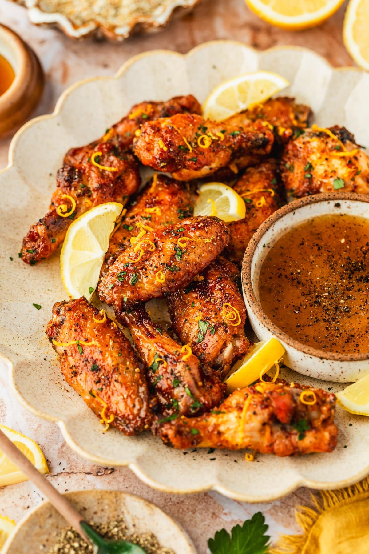A white platter of lemon pepper honey wings and a bowl of sauce on an orange marble counter next to lemons, a yellow linen, and brown bowls of spices, honey, and pepper.