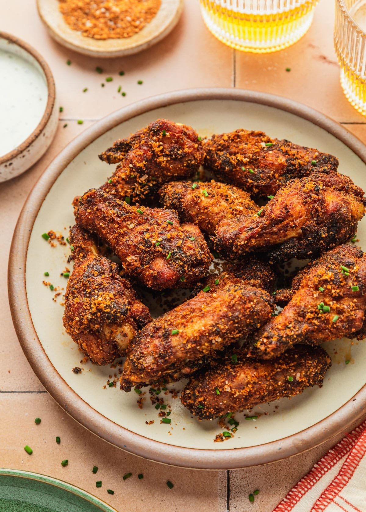 A white stoneware plate of dry rub chicken wings on an orange tiled counter next to a white bowl of ranch, glasses of beer, and orange striped linen.