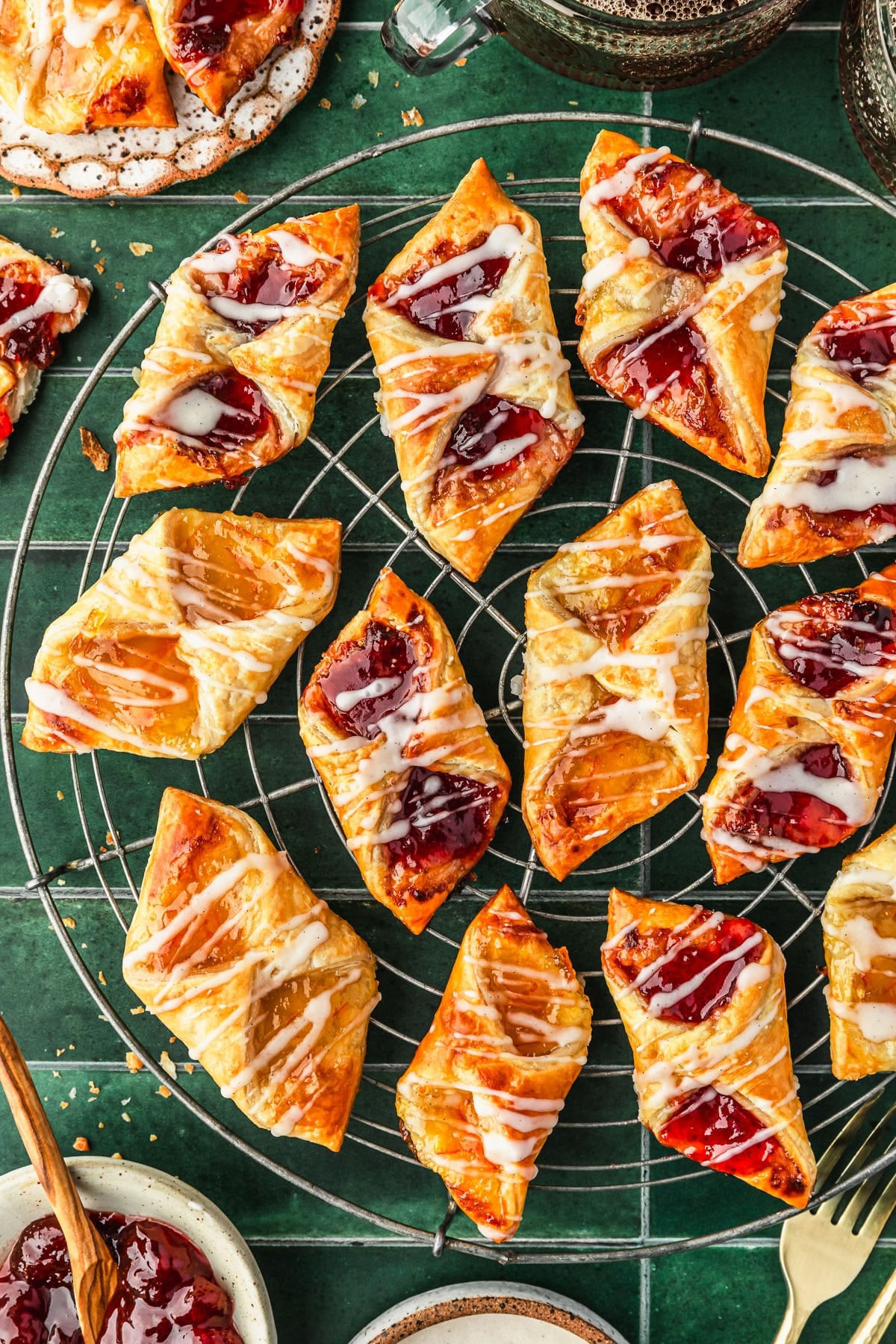 A wire rack of puff pastry jam tarts on a green tiled counter next to cups of coffee, a white plate of tarts, gold forks, and a white bowl of strawberry jam.