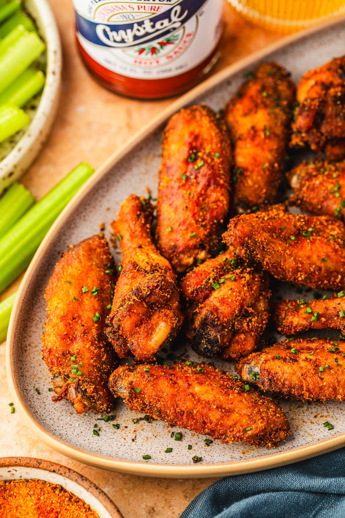 A tan platter of crispy Cajun chicken wings on an orange table next to glasses of beer, a blue linen, a plate of celery, and a bottle of hot sauce.
