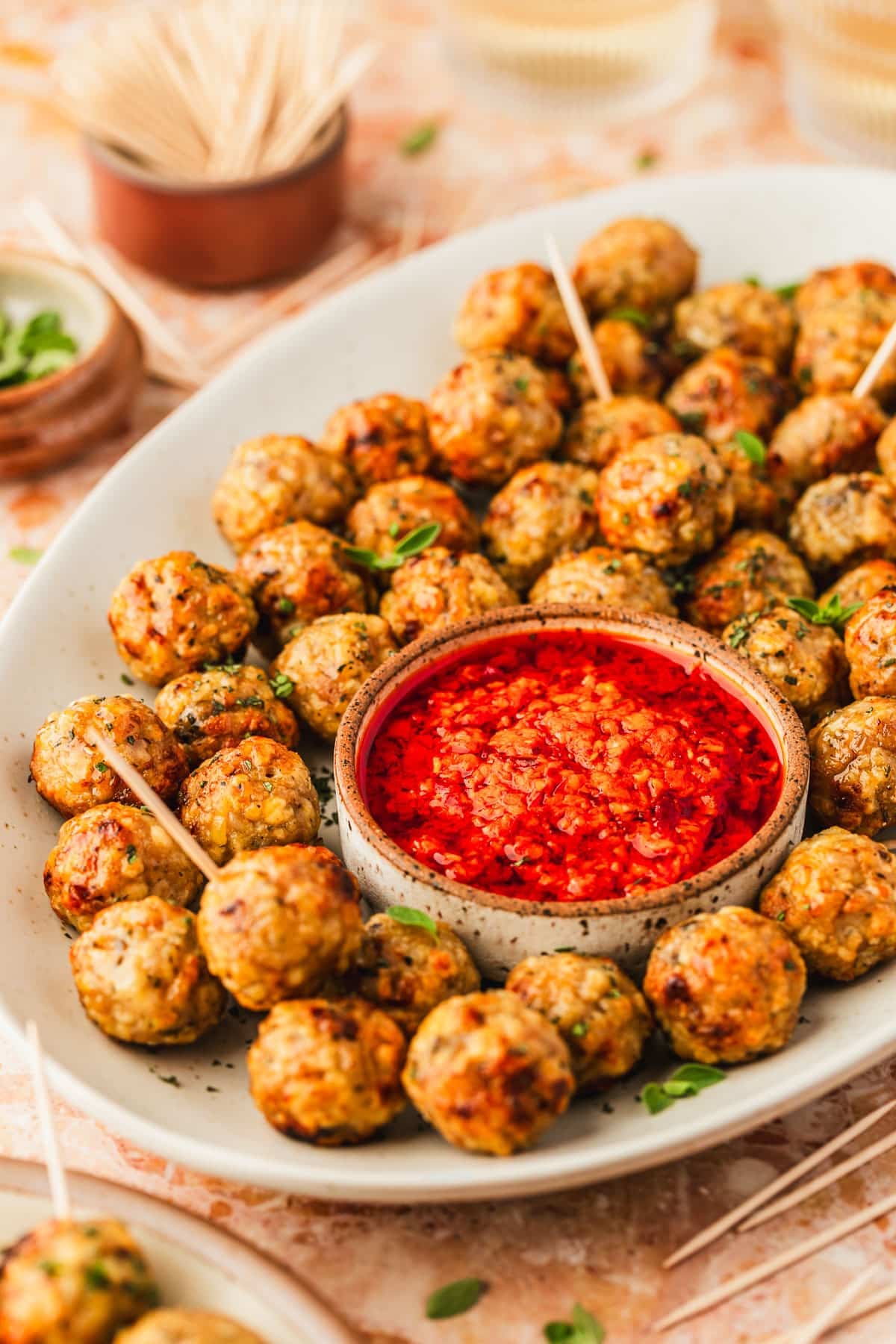 A white plate of air fryer chicken meatballs and a white bowl of red sauce next to glasses of wine, a brown plate of meatballs, and brown bowls of oregano and toothpicks on an orange marble counter.
