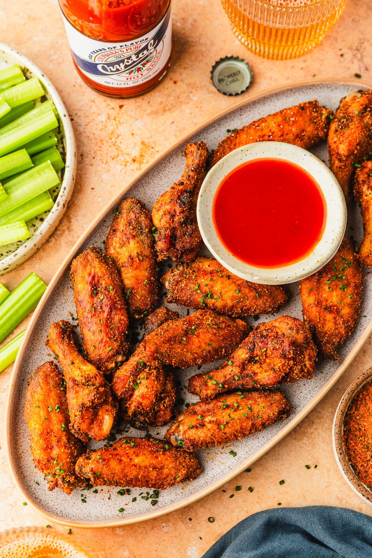 A stoneware platter of Cajun chicken wings and a white bowl of hot sauce on an orange counter next to glasses of beer, a bottle of hot sauce, a plate of celery, and a blue napkin.