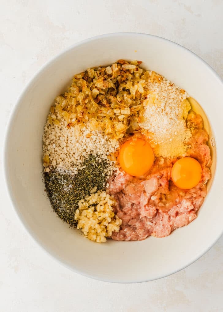 A white bowl of ground meat, breadcrumbs, parmesan, herbs, onions, and eggs on a white counter.