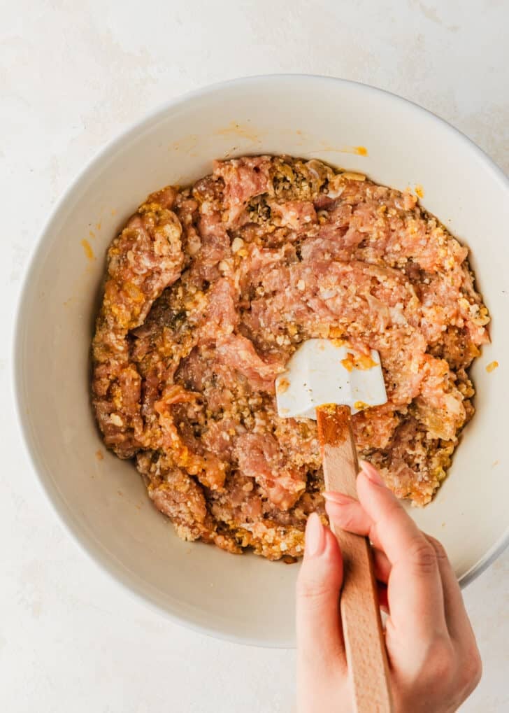 A hand using a rubber spatula to mix meatball mixture in a white bowl.