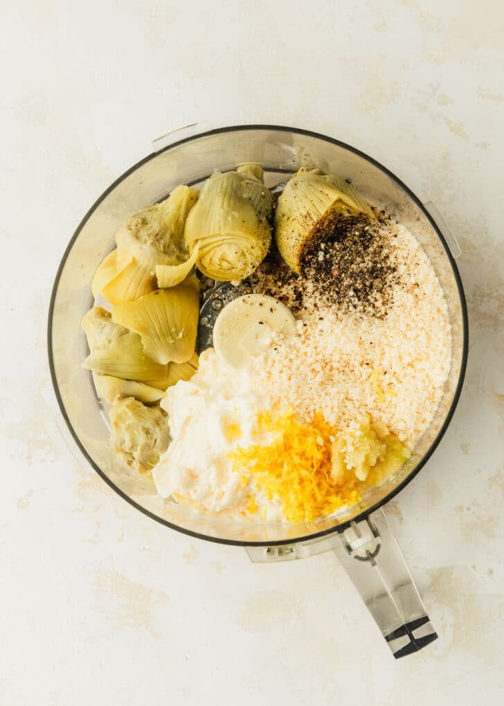 A food processor of artichoke hearts, mayo, parmesan, lemon zest, salt, and pepper on a tan table.