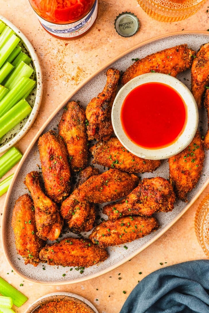 A stoneware plate of Cajun chicken wings and a white bowl of hot sauce on an orange stone counter next to glasses of beer, a bottle of Louisiana hot sauce, a plate of celery, and a blue linen.