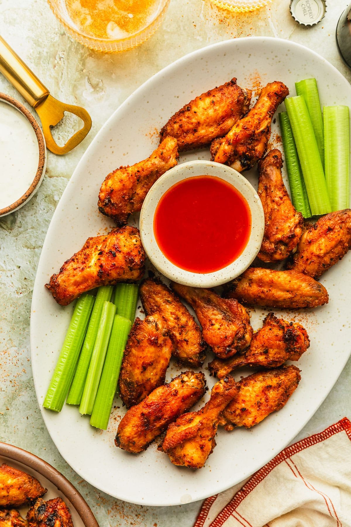 A white platter of crispy oven-baked chicken wings, celery sticks, and a white bowl of hot sauce next to a plate of wings, orange and cream striped linen, glasses of beer, and brown bowl of ranch with a green marble background.