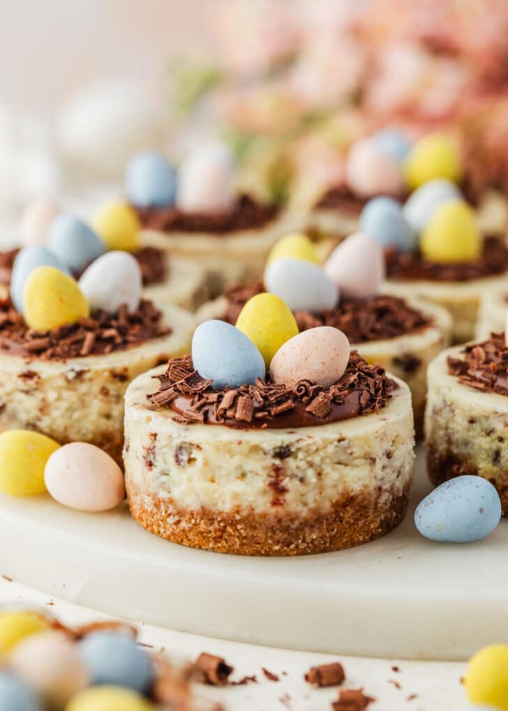 A white platter of mini Easter egg cheesecakes on a tan counter next to pink flowers, a brown bowl of candies, and white eggs.