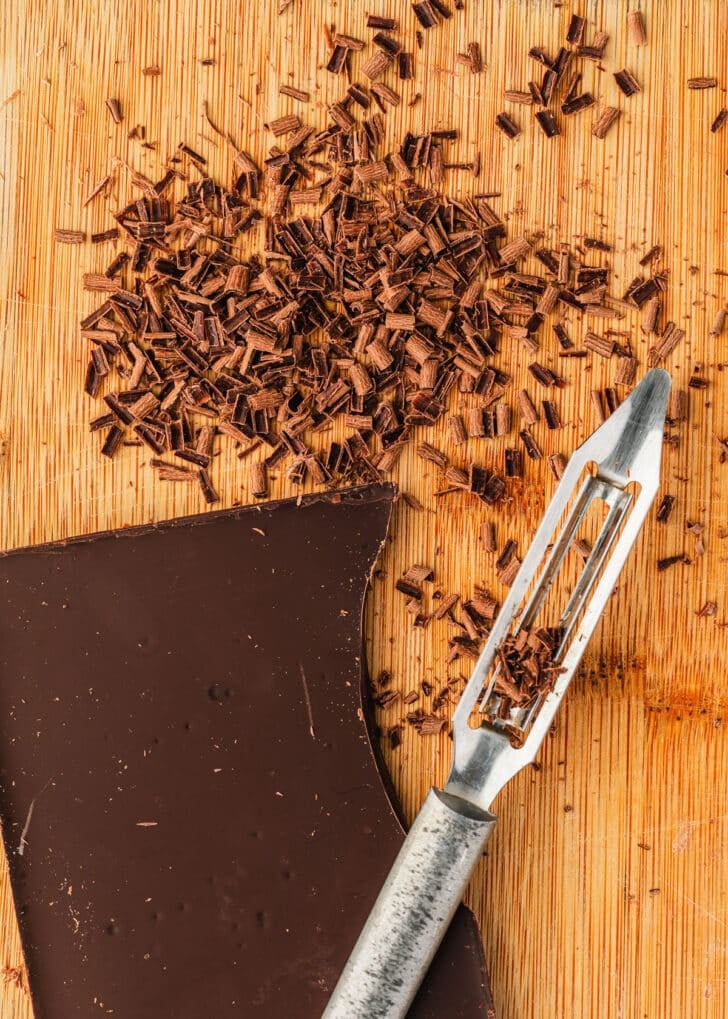 A bar of chocolate, vegetable peeler, and chocolate shavings on a wood board.