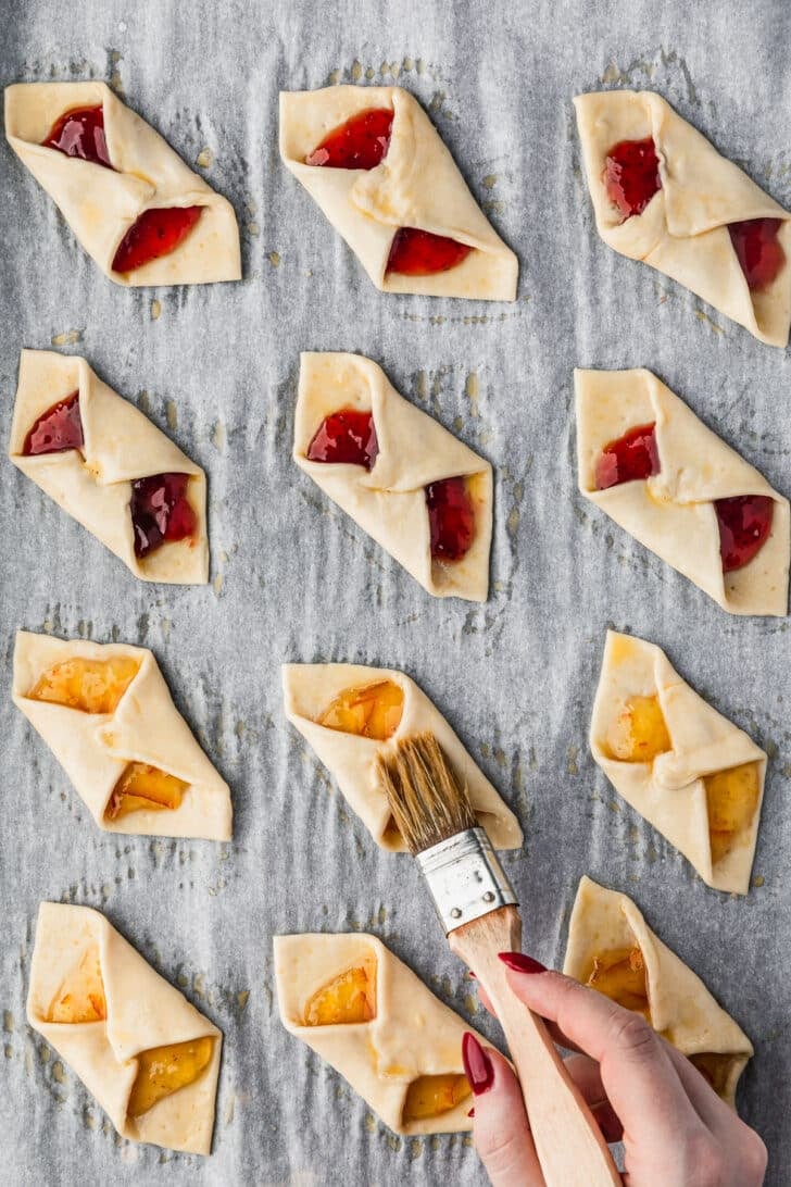 A hand using a pastry brush to brush puff pastry jam tarts with egg wash.