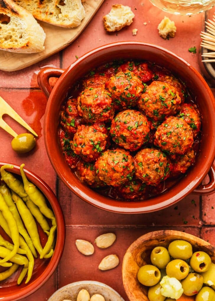 Red bowls of Spanish meatballs and peppers next to a wood board of bread, wood bowl of olives, brown bowl of almonds, and glass of white wine on a terracotta tiled table.