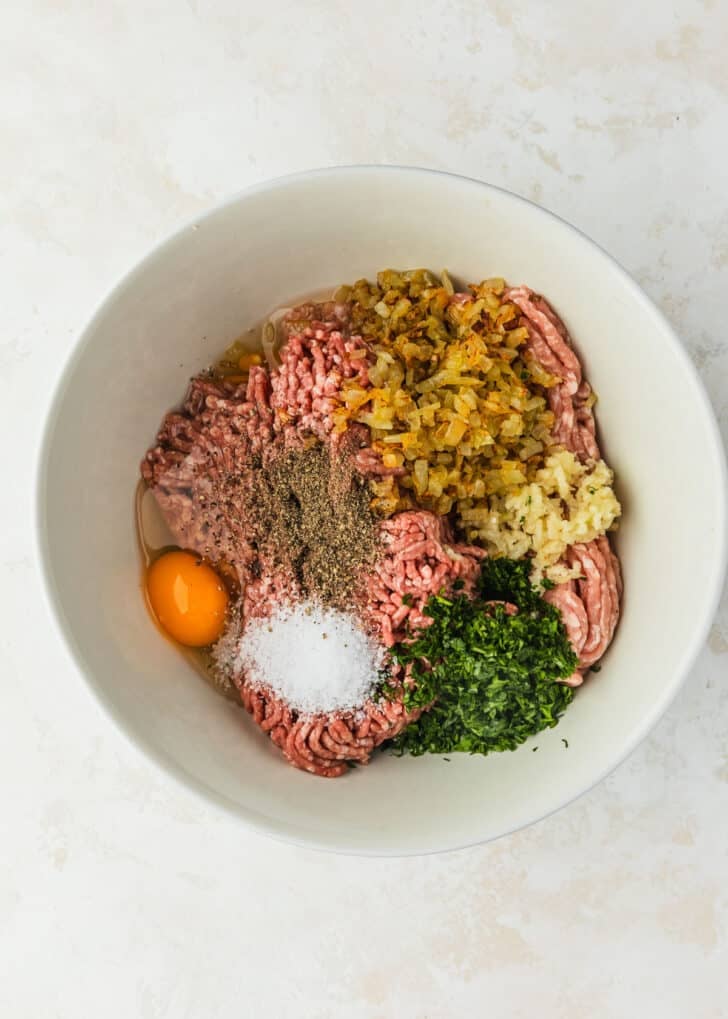 A white bowl of ingredients for Spanish meatballs on a beige table.