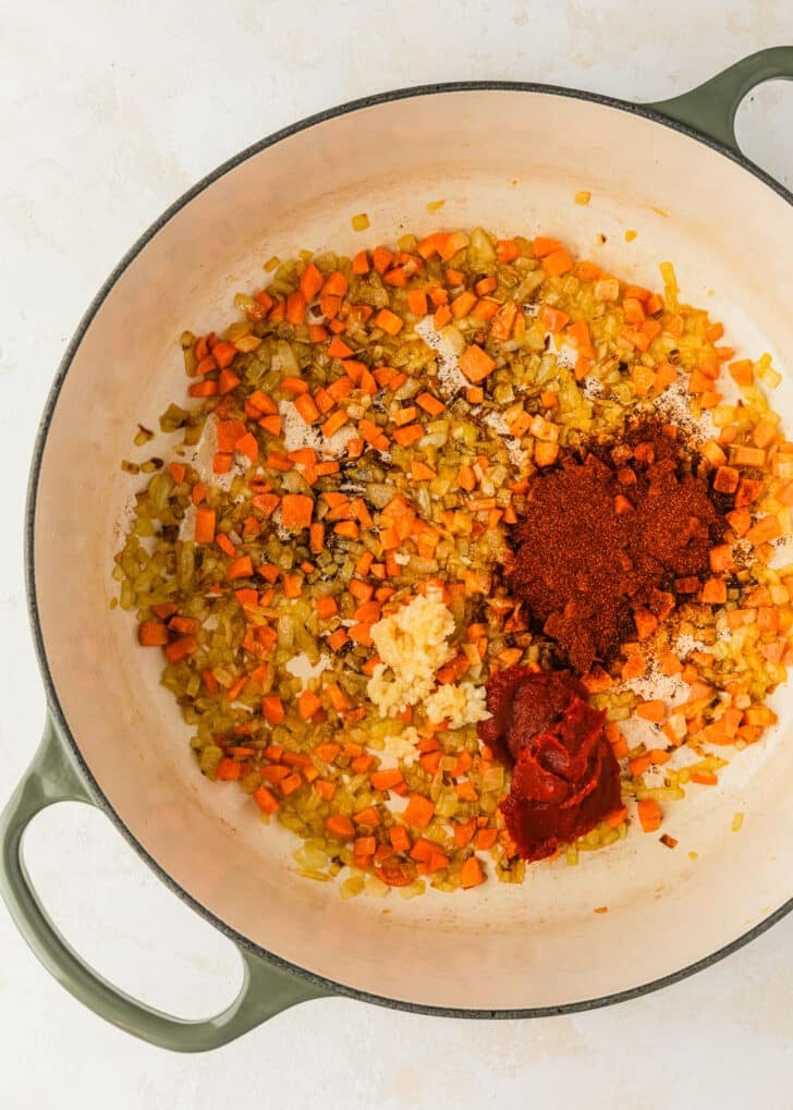 A white pot of onions, carrots, garlic, spices, and tomato sauce on a beige table.