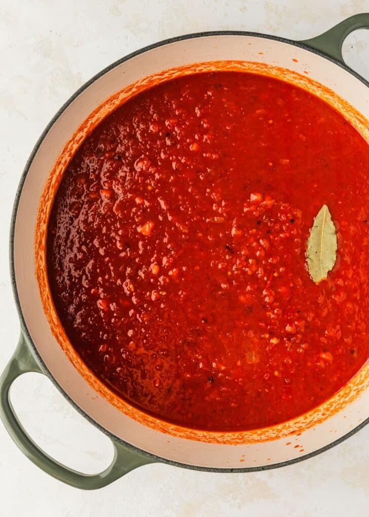 A white pot of tomato sauce with a bay leaf on a tan counter.