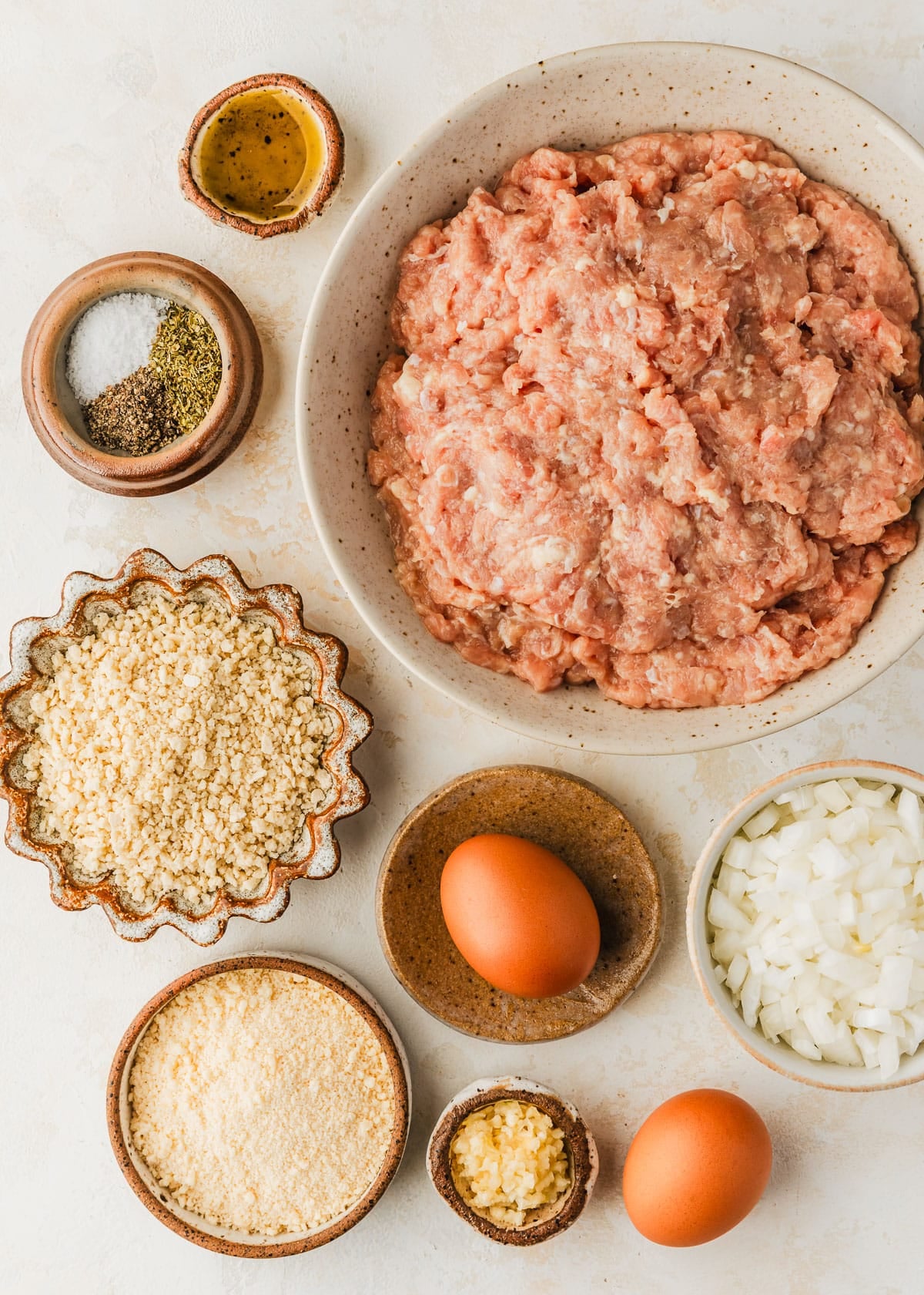 White and brown bowls of ground meat, spices, breadcrumbs, parmesan, onion, eggs, garlic, and oil on a tan counter