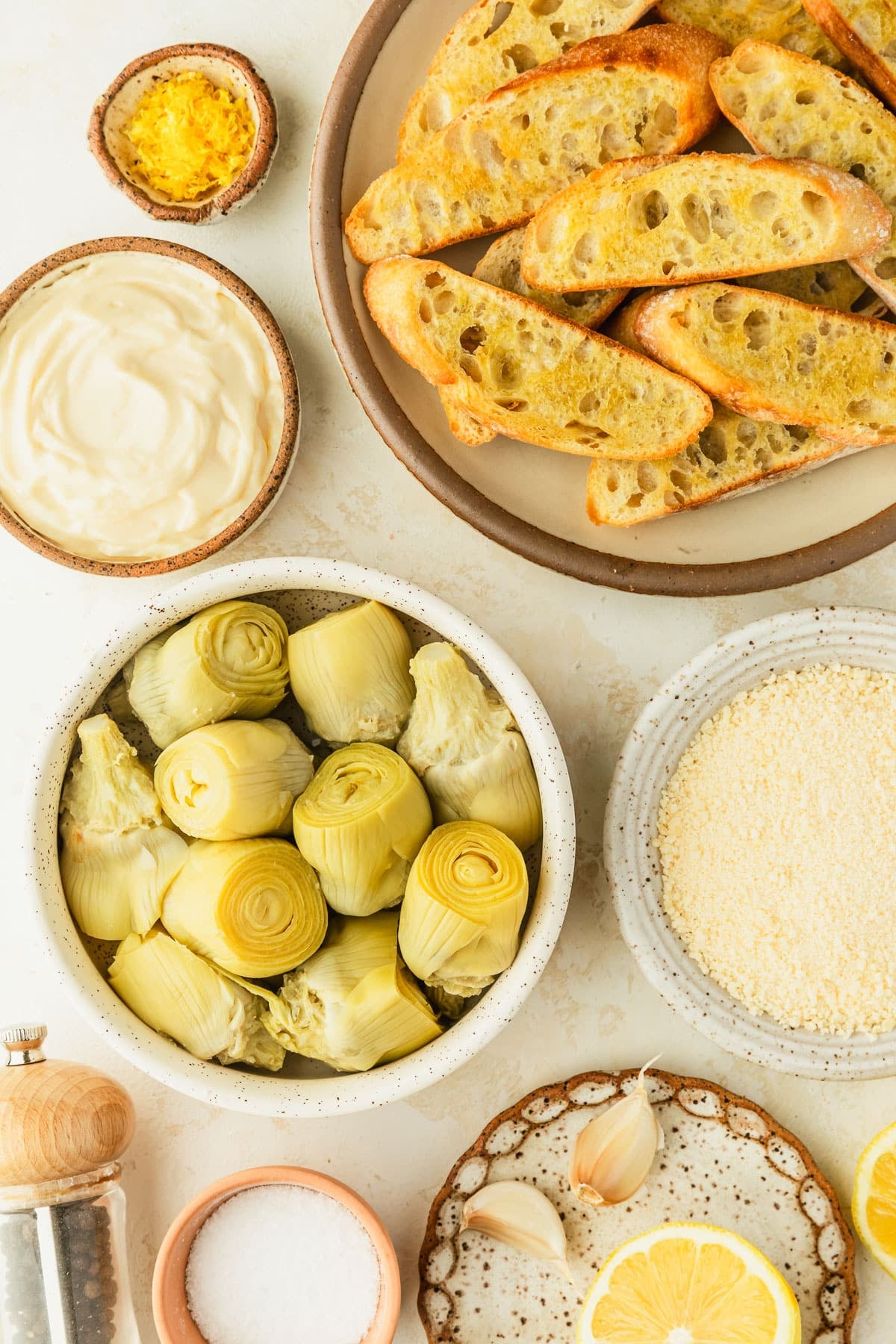 White and brown bowls of bread, lemon zest, mayo, parmesan, artichoke hearts, salt, garlic, and lemons on a beige counter.