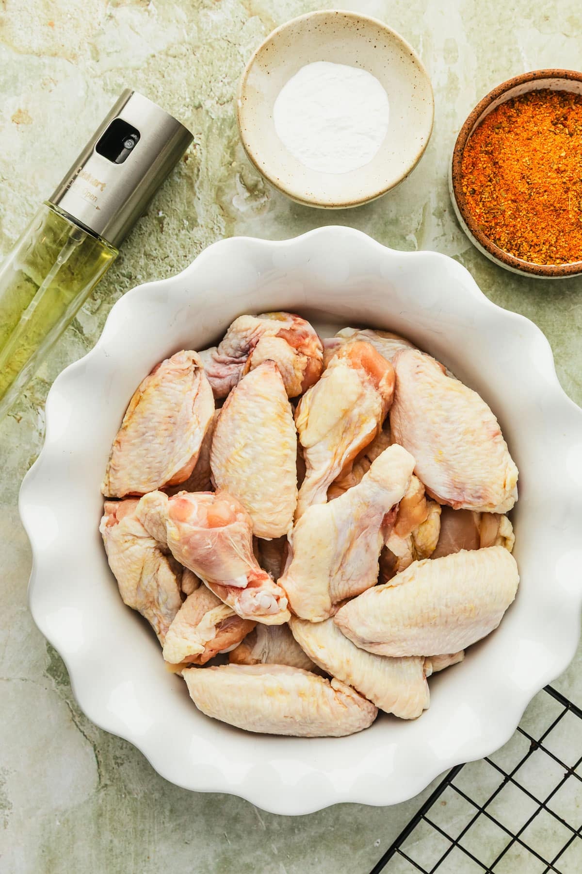 White and brown bowls of chicken wings, baking powder, and spices on a green marble counter next to cooking spray and a wire rack.
