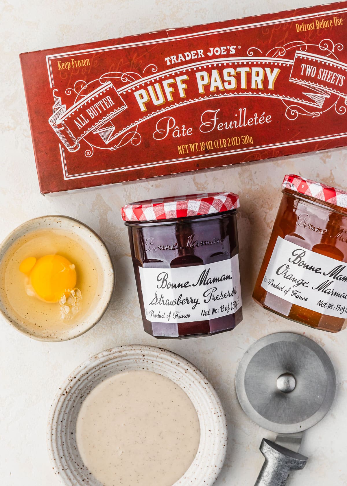 A box of pastry, jars of preserves, and white bowls of glaze and an egg on a tan counter.