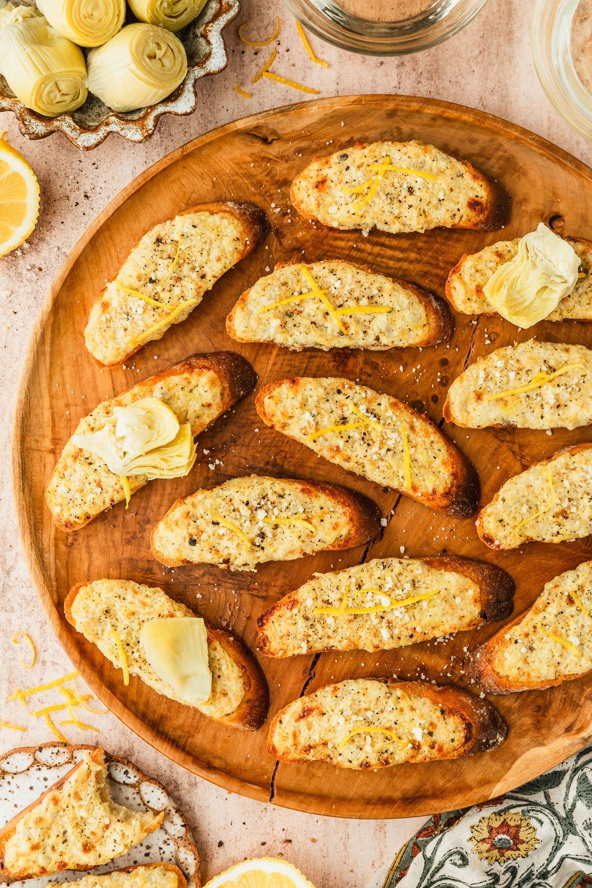 A wood platter of artichoke bruschetta with parmesan and lemon zest on a brown stone counter next to glasses of white wine, a floral linen, sliced lemons, and a brown bowl of artichokes.