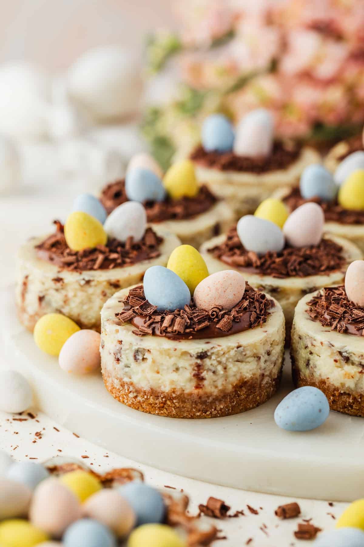A marble platter of mini egg cheesecakes with ganache nests next to a brown bowl of candies, pink flowers, and white ceramic bunnies on a white counter.