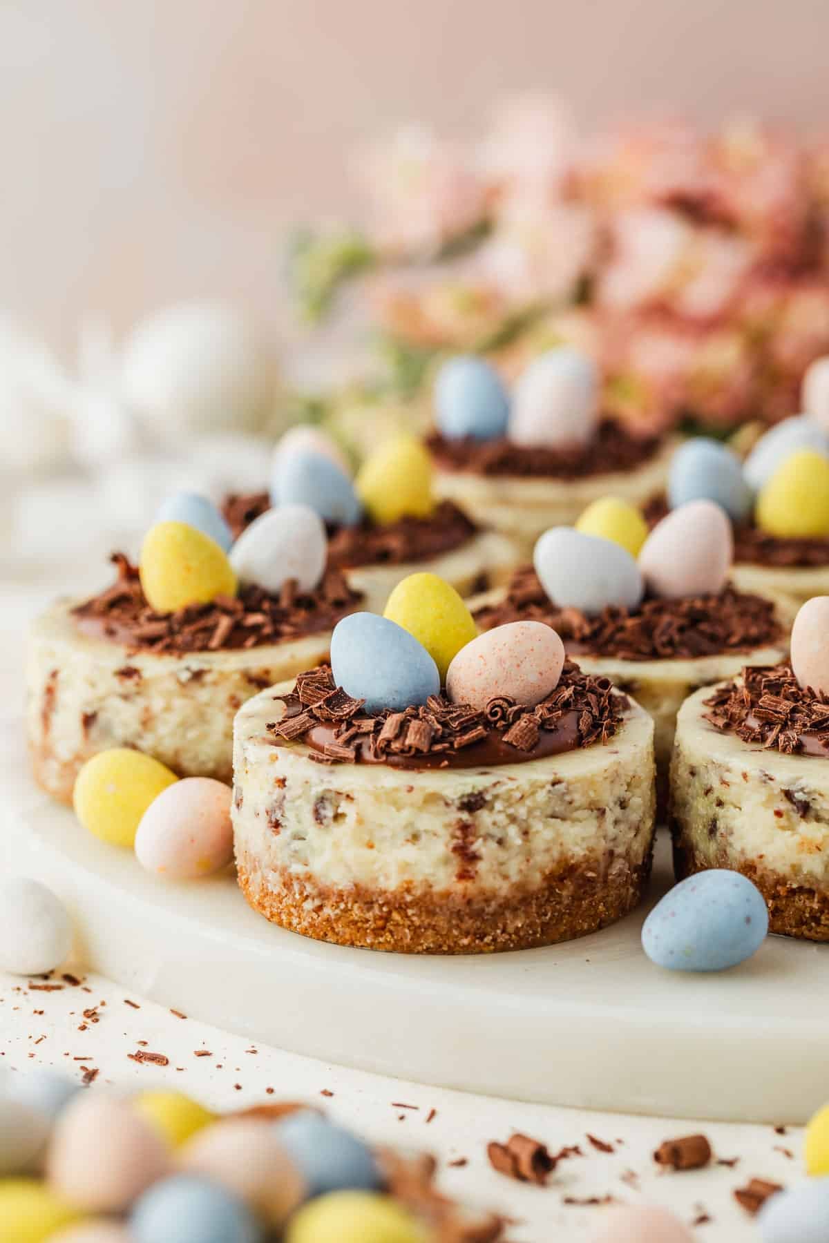 A white platter of mini Easter egg cheesecakes next to a bowl of mini egg candies and pink flowers on a white backdrop.
