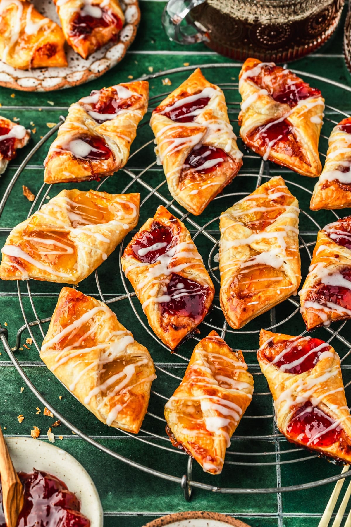 A wire rack of jam puff pastry tarts on a green tiled counter next to a white plate of tarts, cups of coffee, and a white bowl of jam.