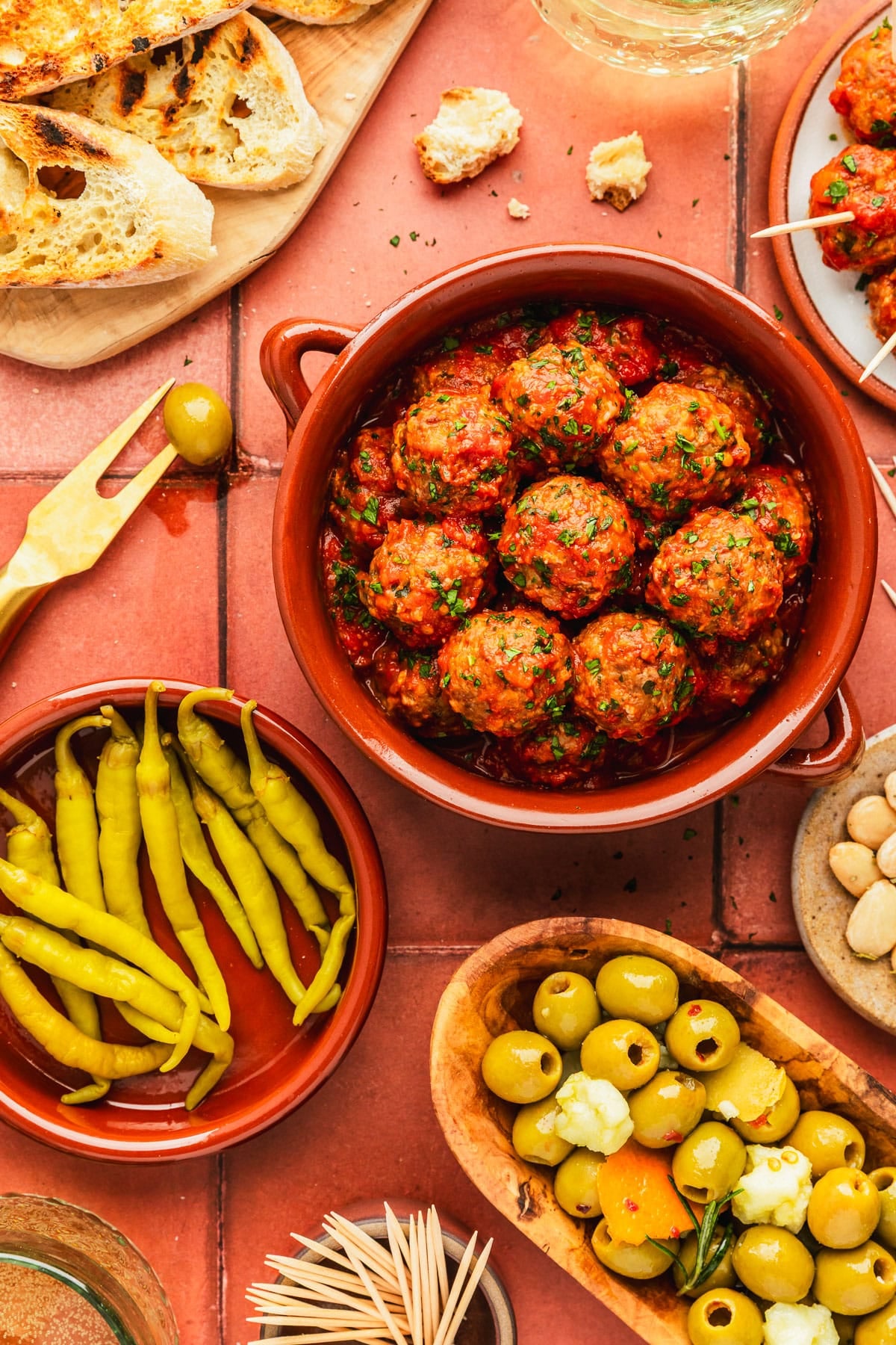 A red bowl of Spanish meatballs tapas next to a red bowl of peppers, a wood board of bread, a wood bowl of olives, a tan bowl of almonds, and glasses of white wine on a red tile counter.