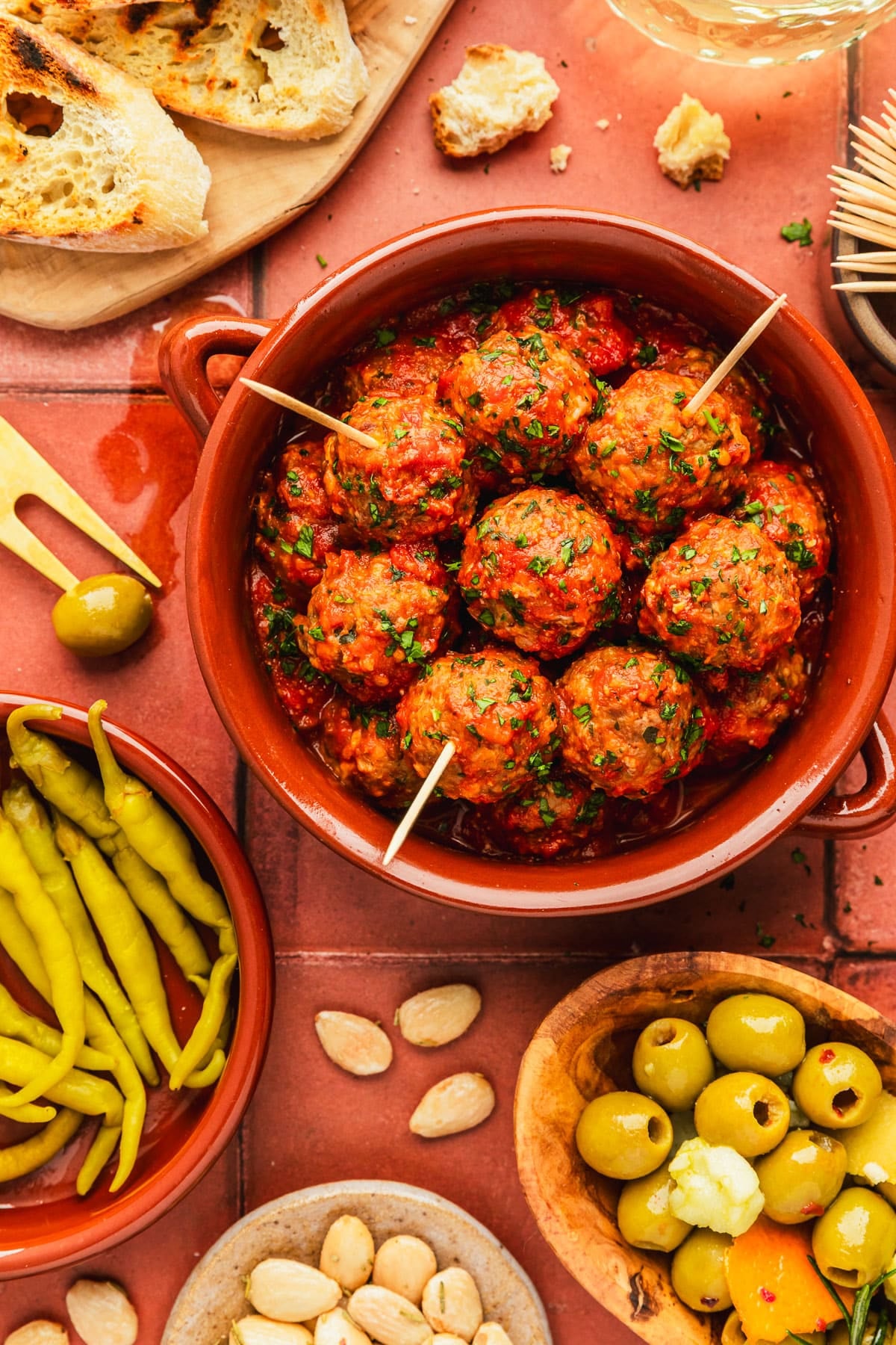 Red bowls of tapas-style Spanish meatballs and peppers on a terracotta tiled counter next to wood bowls of bread and olives, glasses of white wine, and a brown bowl of almonds.