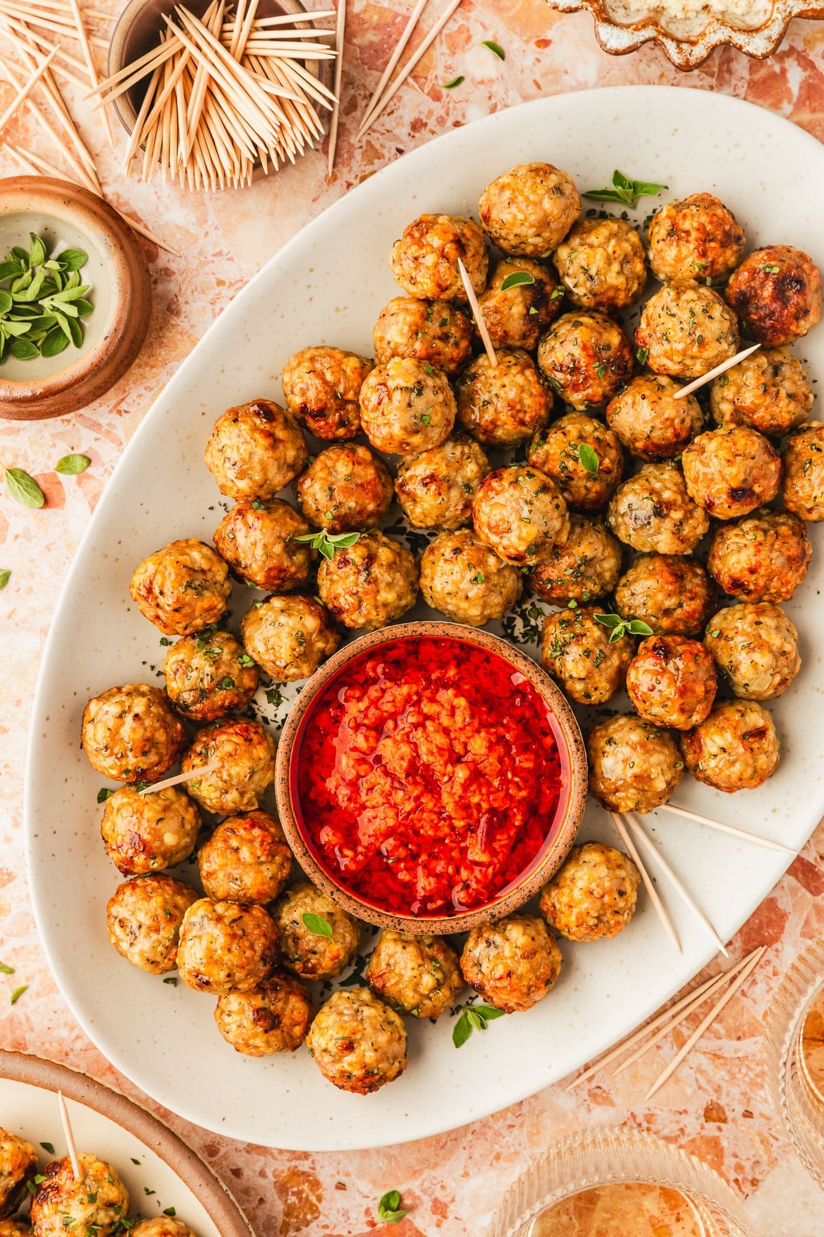 A white platter of air fryer chicken meatballs with a bowl of red sauce next to glasses of wine, a stoneware plate of meatballs, and brown bowls of oregano and toothpicks on an orange stone counter.