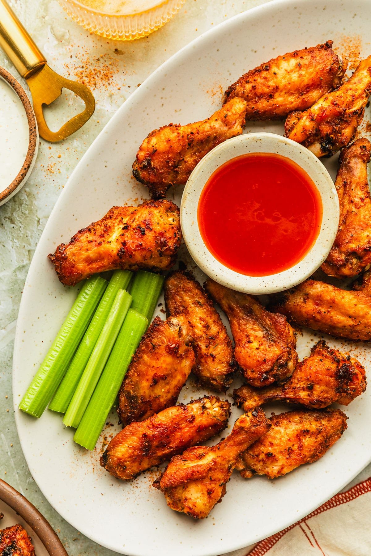 A white platter of crispy oven-baked chicken wings, celery, and a white bowl of hot sauce on a green marble counter next to a brown bowl of ranch, glasses of beer, and a gold bottle opener.
