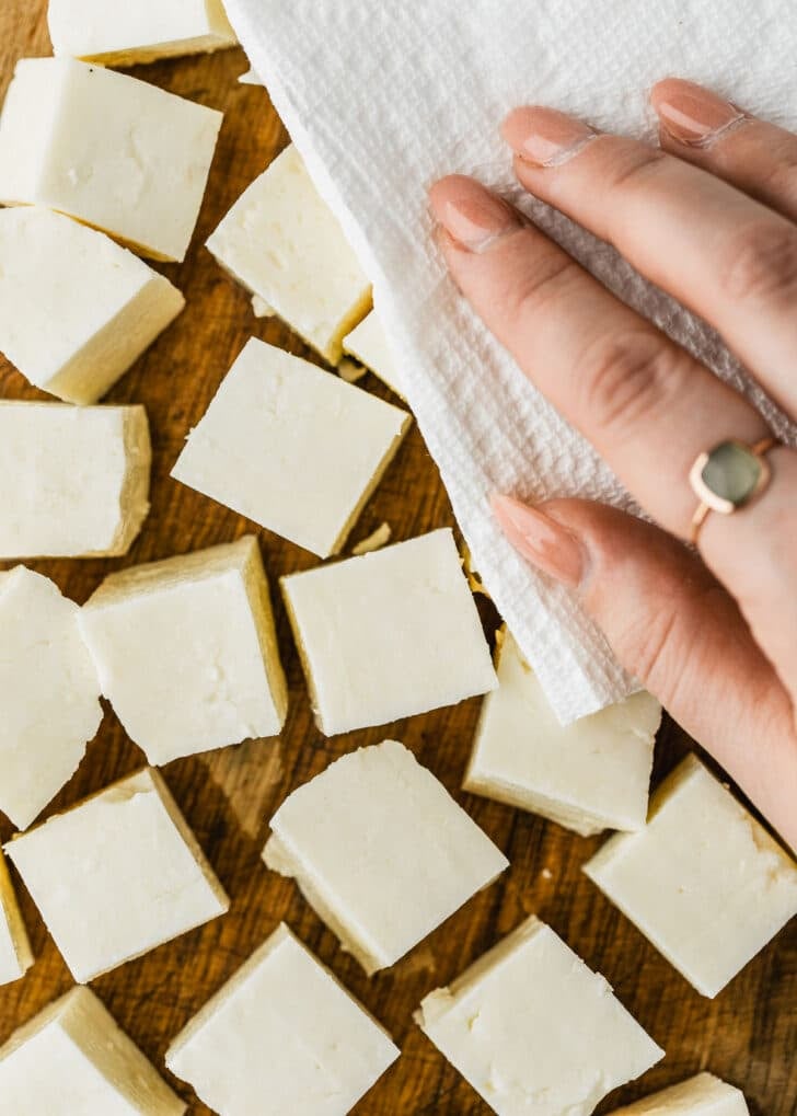 A hand using paper towels to dry halloumi cubes on a wood board.