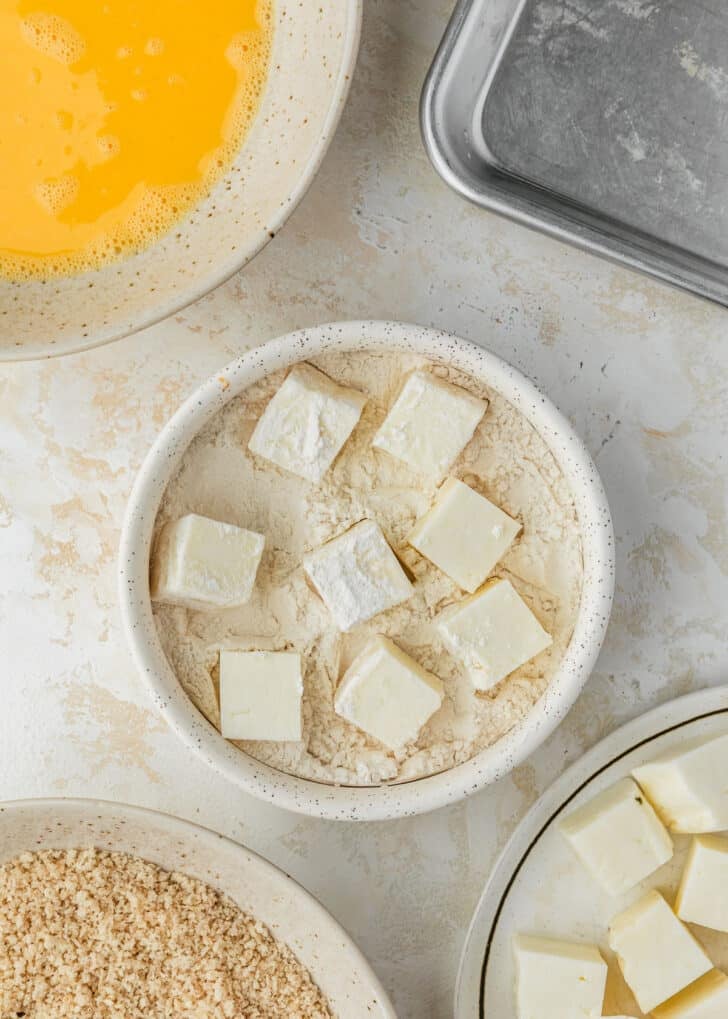 A white bowl of flour with cheese cubes next to white bowls of cheese, breadcrumbs, and whisked eggs on a tan counter.