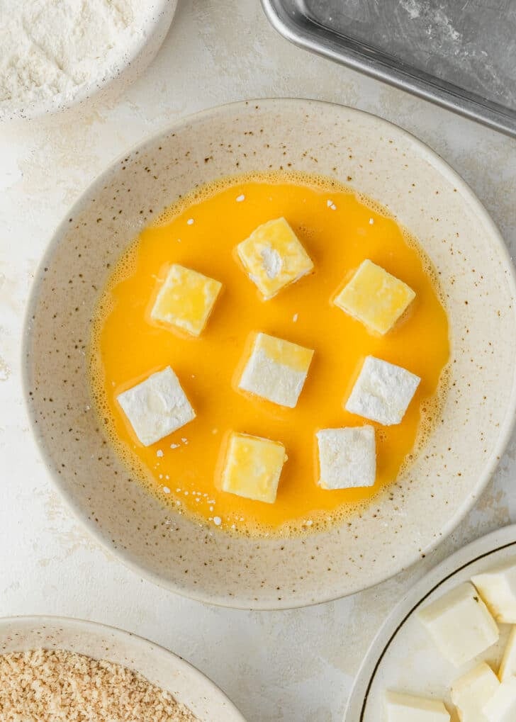 Cubes of halloumi in a white bowl of whisked eggs next to a white bowl of flour, white bowl of breadcrumbs, and sheet pan on a beige table.