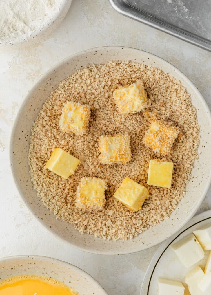 Raw halloumi bites in a white bowl of breadcrumbs next to white bowls of cheese, eggs, and flour on a tan table.