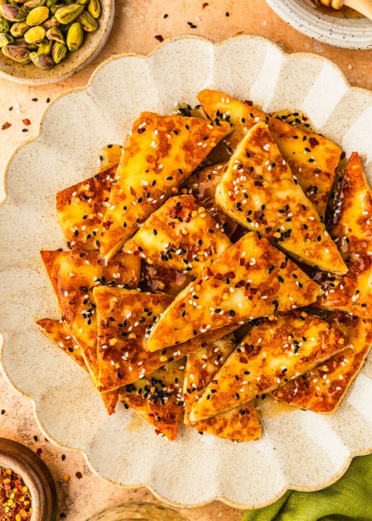 A white platter of hot honey halloumi with sesame seeds next to a white bowl of honey, brown bowls of pepper flakes and pistachios, and an olive green linen on an orange table.