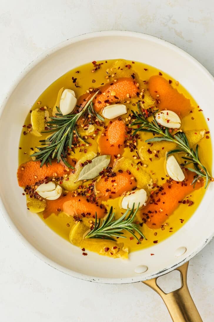 A white pan of olive oil, orange peels, lemon peels, garlic, and rosemary sprigs on a tan counter.