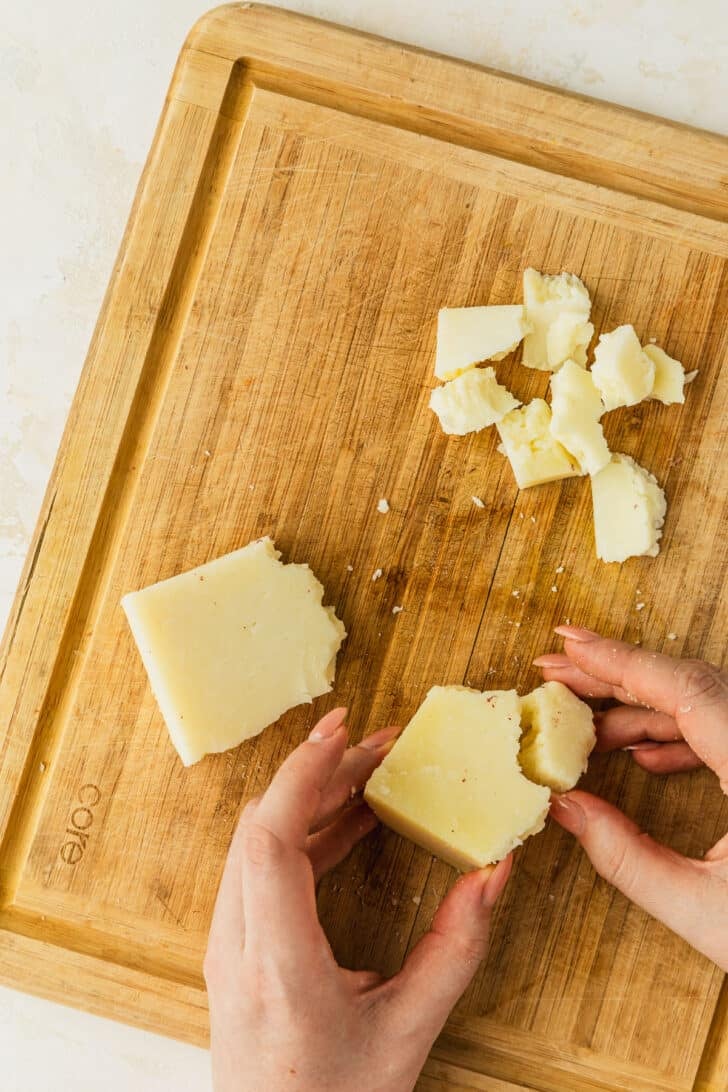Hands breaking a block of Manchego into shards on a wood board.