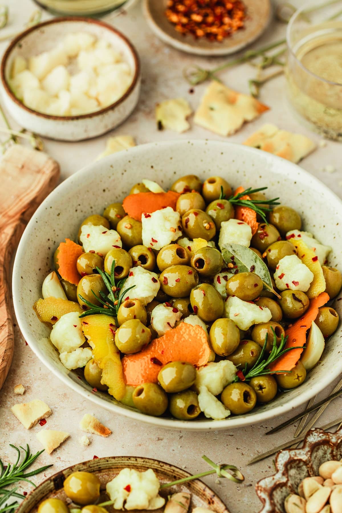 A white bowl of marinated Spanish olives with manchego, orange peel, lemon peel, garlic, and rosemary next to a wood board of crackers, glasses of cider, and brown bowls of olives, pepper flakes, and cheese on a brown stone counter.