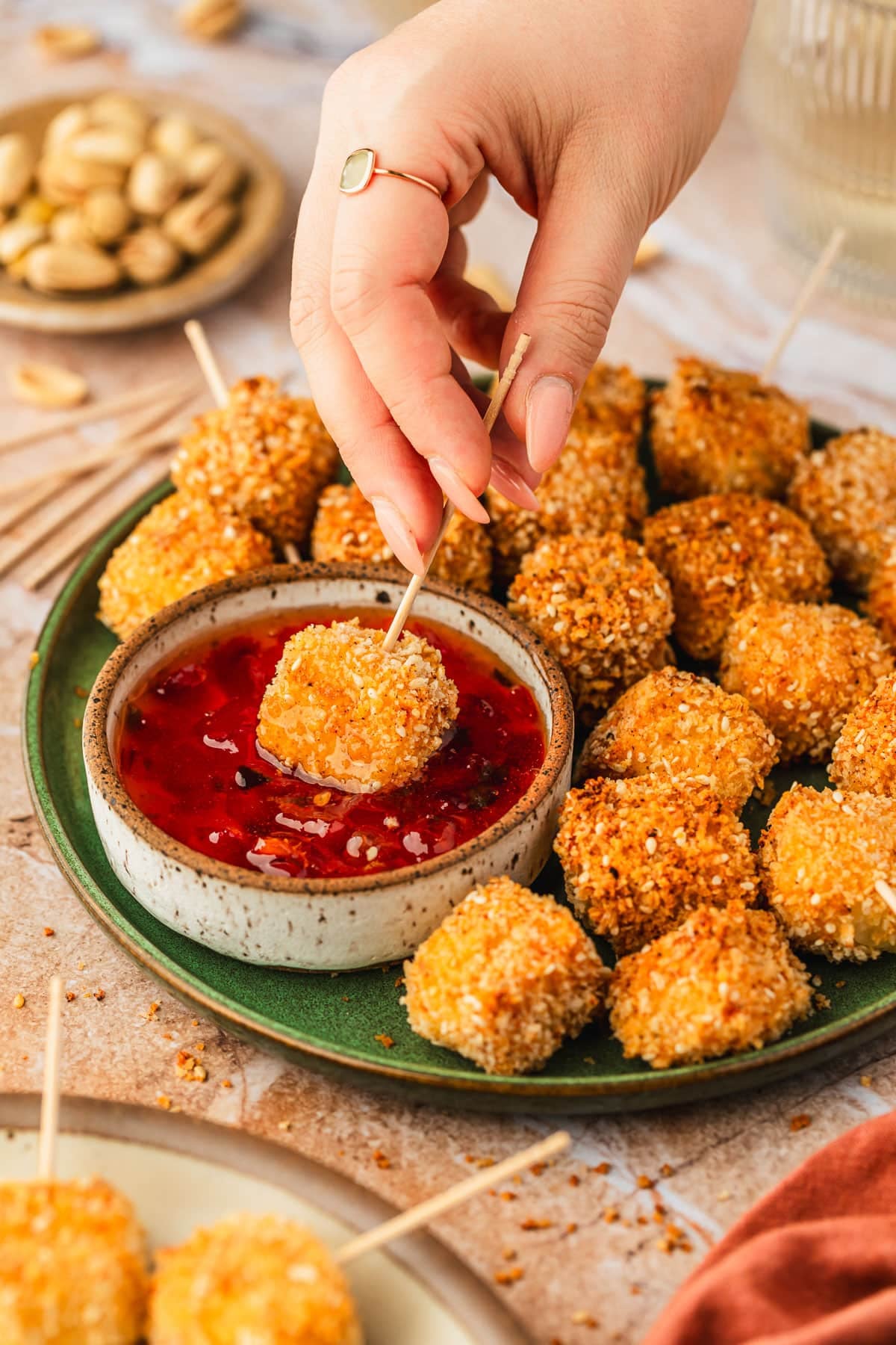 A hand dipping a cheese bite into a white bowl of red pepper jelly on a green platter of crispy halloumi bites next to an orange linen, glass of white wine, and brown bowl of pistachios on a tan marble table.