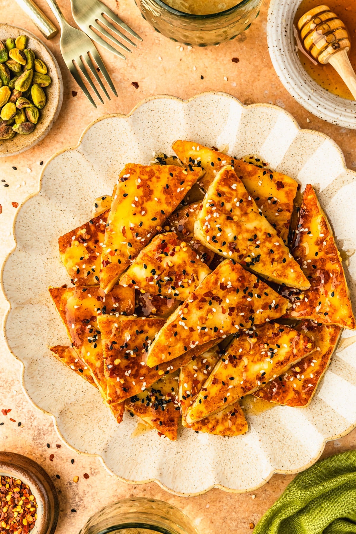 A white platter of hot honey halloumi next to glasses of white wine, a white bowl of hot honey, brown bowls of pistachios and pepper flakes, and an olive green linen on an orange background.