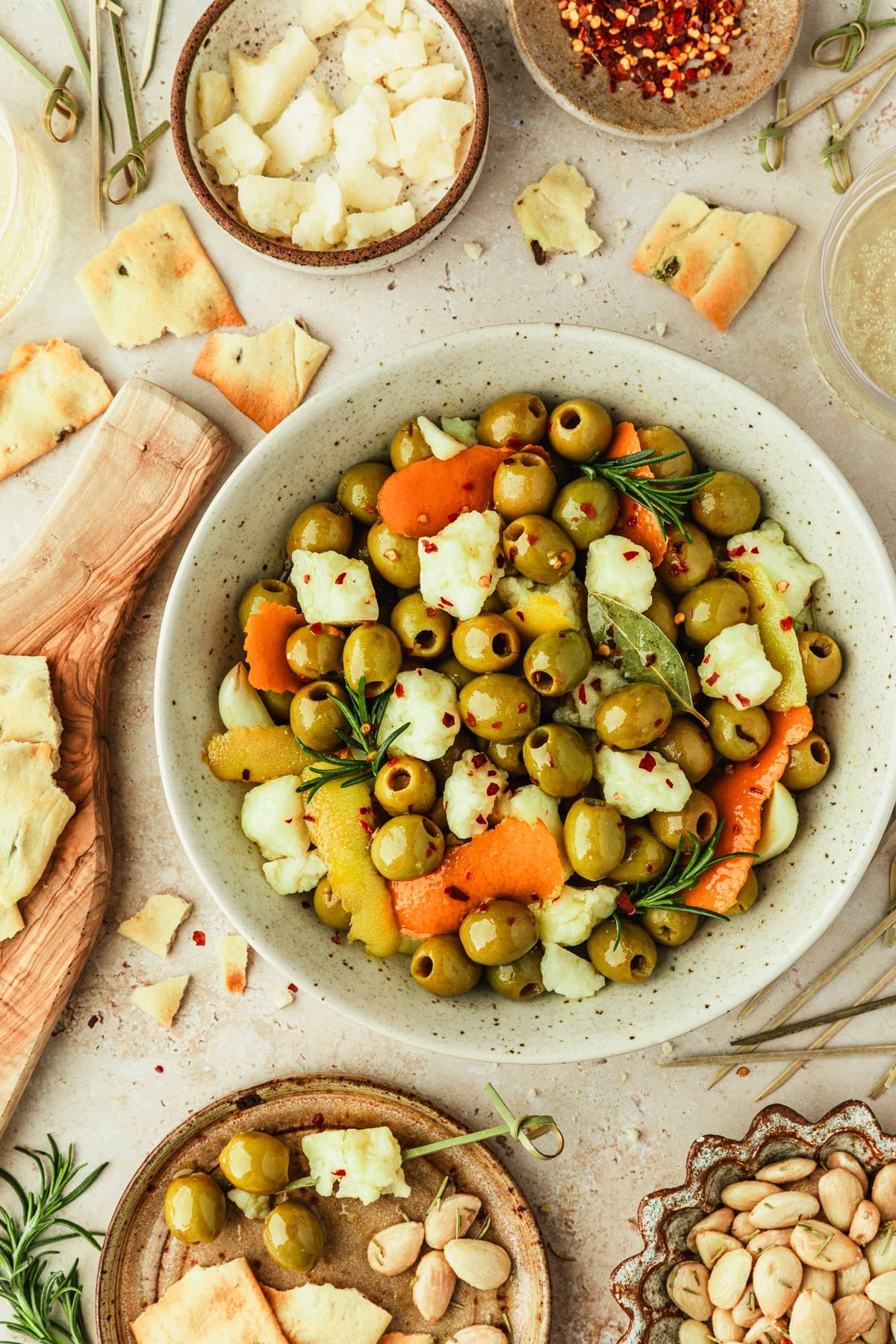 A white bowl of marinated Spanish olives and manchego on a tan counter next to glasses of cider, a wood board of crackers, and brown bowls of almonds, cheese, and pepper flakes.