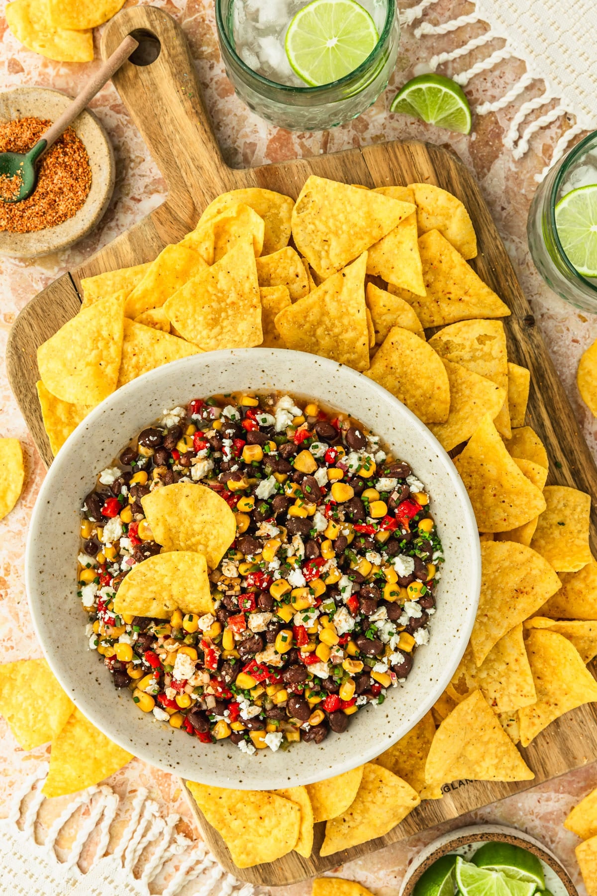 A white bowl of black bean and corn dip with feta on a wood board of corn chips next to glasses of ranch water, white napkins, and brown bowls of limes and Tajin on an orange stone table.