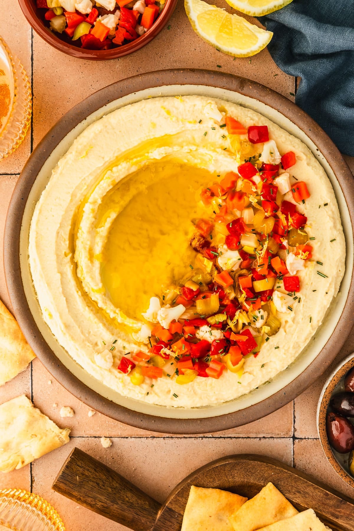 A tan platter of butter bean dip with olive oil and giardiniera next to glasses of beer, a blue linen, a wood board of crackers, and a brown bowl of olives on an orange-brown tiled counter.