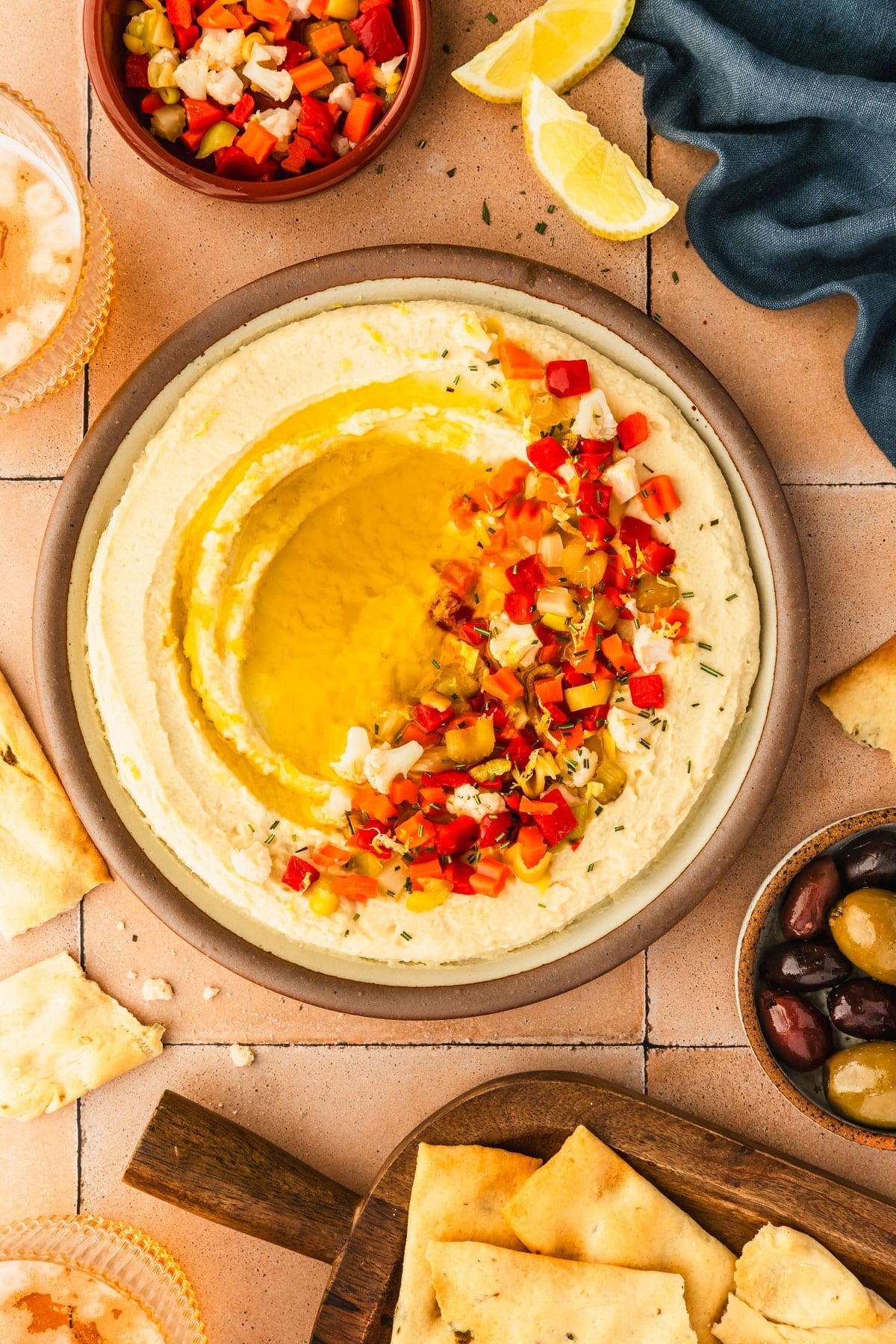 A beige platter of butter bean dip with giardiniera next to a blue linen, glasses of beer, a wood tray of crackers, and a brown bowl of olives on a terracotta tile counter.
